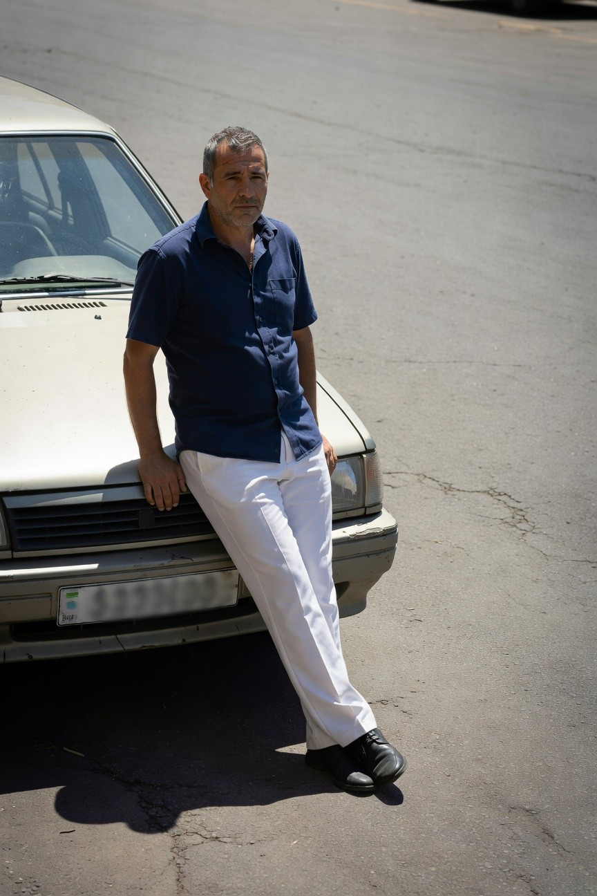 Middle-aged man with short graying hair leans casually against a beige sedan on a sunny urban street, dressed in a short-sleeve navy blue button-up shirt tucked into slim white trousers and black leather dress shoes, evoking relaxed summer sharpness