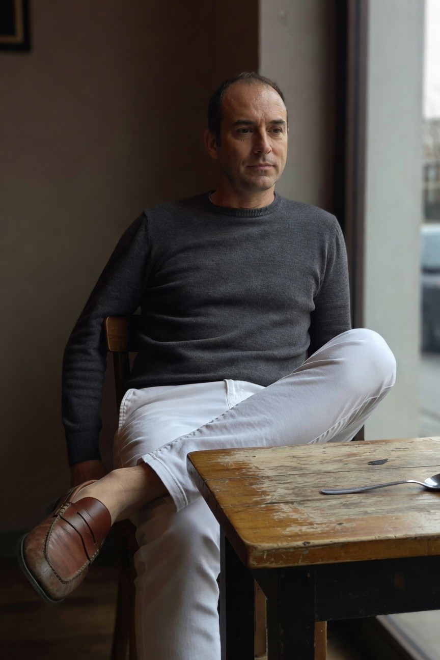 Middle-aged man in gray crewneck sweater, slim white trousers, and brown woven loafers, seated casually by a cafe window with legs crossed over a wooden table