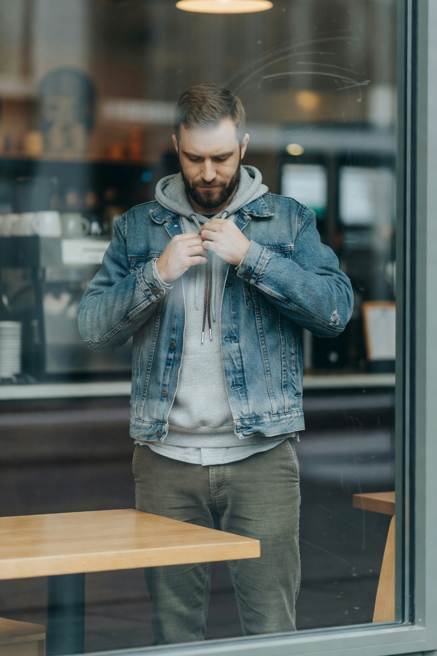 Man buttoning a light wash distressed denim jacket over a gray hoodie with drawstrings, paired with straight-leg khaki pants, standing inside near a cafe window