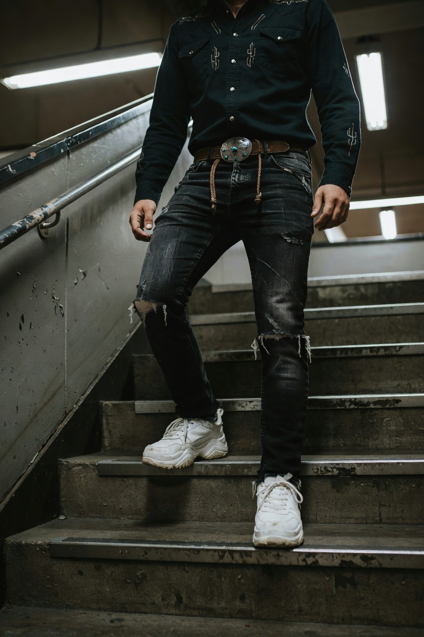 Man in navy embroidered western shirt, large turquoise belt buckle, ripped black slim jeans, and white chunky sneakers, posing mid-stride on concrete stairs in urban setting
