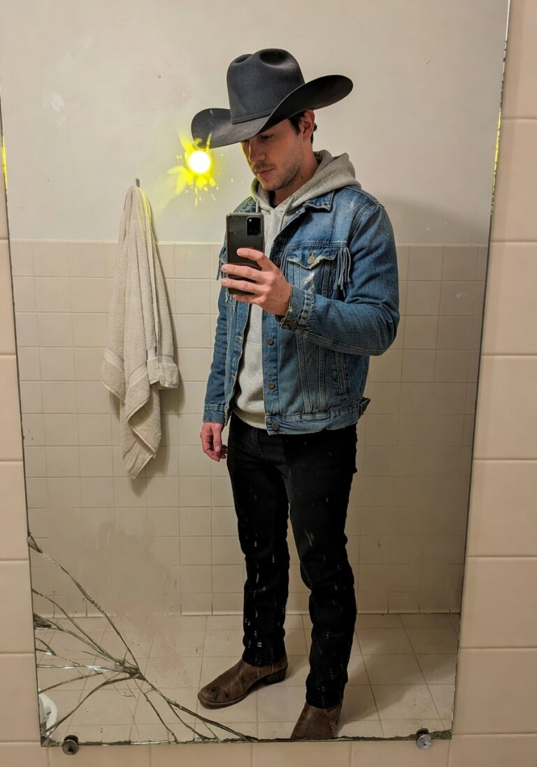 Man taking mirror selfie in bathroom wearing dark cowboy hat, frayed light blue denim jacket over gray hoodie, dark jeans, and tan cowboy boots