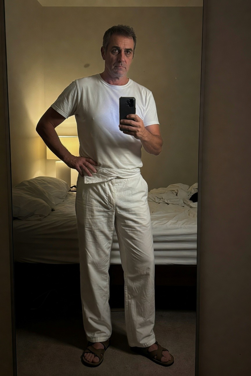 Middle-aged man with salt-and-pepper hair stands confidently in a dimly lit bedroom mirror selfie, wearing a fitted white t-shirt, loose white parachute pants with drawstring waist, and brown leather sandals, hand on hip holding phone, white bedding and lamp in background