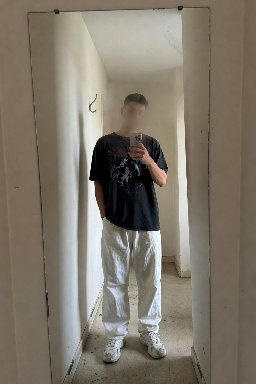 A man takes a mirror selfie in a narrow beige hallway, wearing a black graphic band t-shirt, baggy white parachute pants, and white sneakers, hands in pockets for a relaxed streetwear vibe