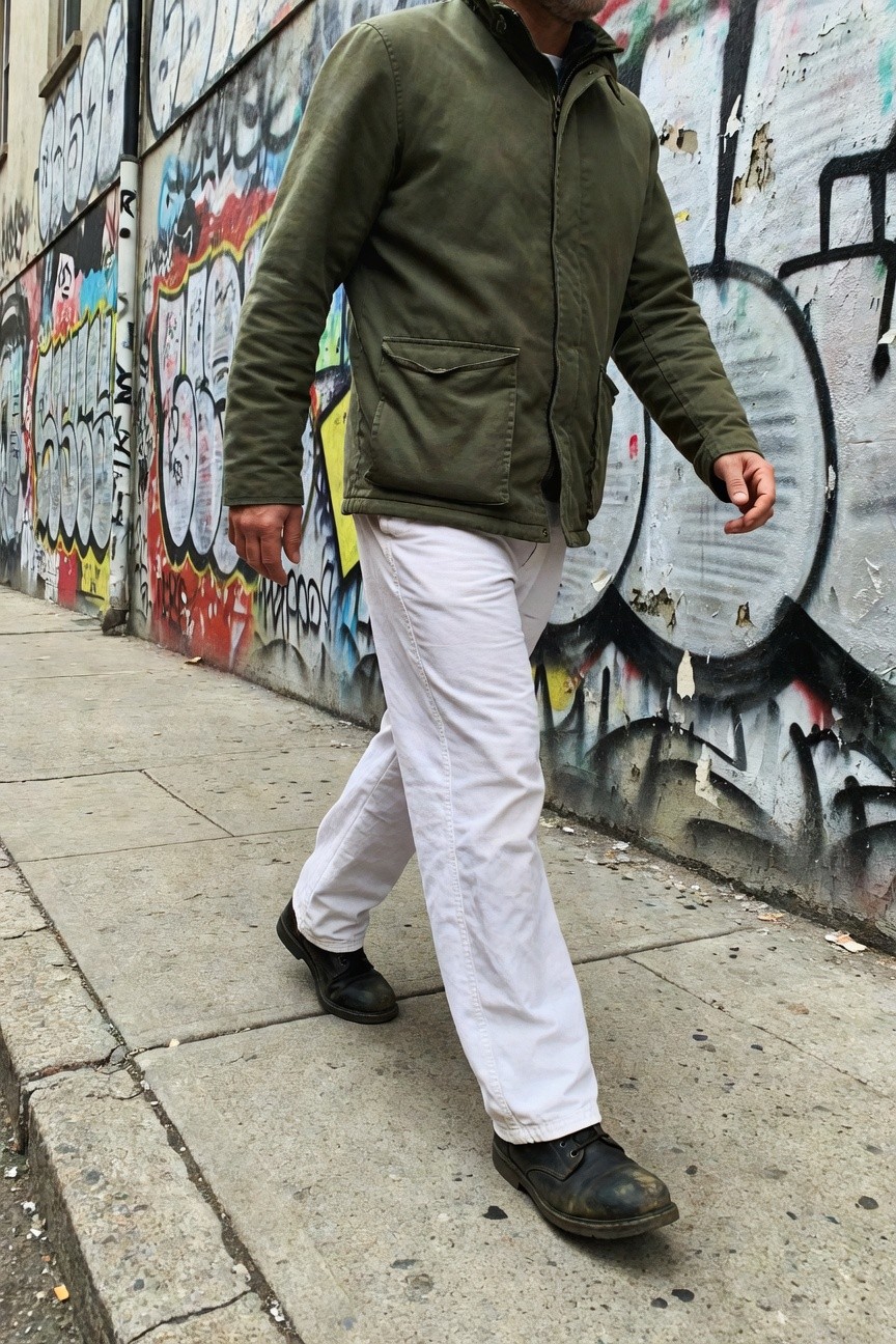 Man in olive green field jacket, white baggy parachute pants, and black boots walking along graffiti-covered urban wall on sidewalk