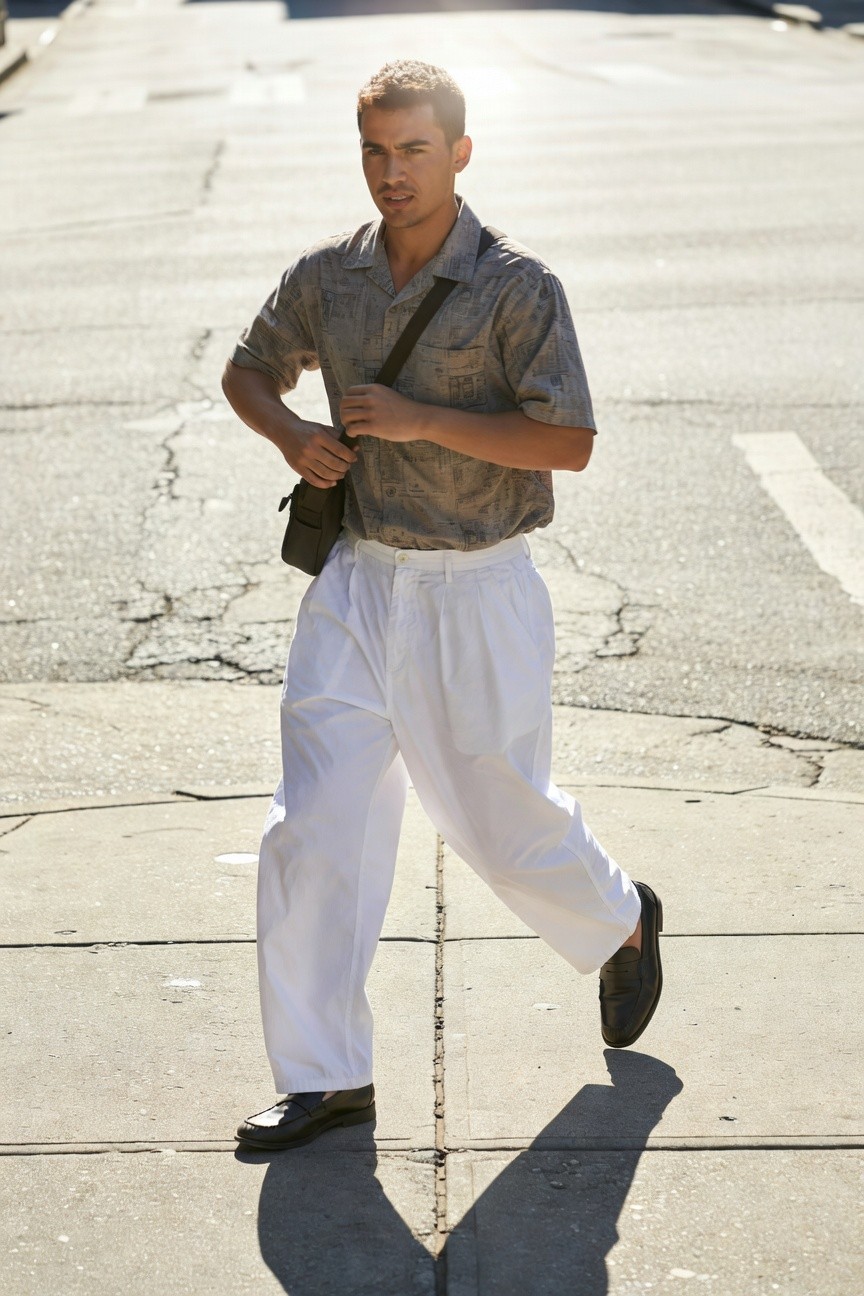 Blond man in short-sleeve gray patterned shirt, white wide-leg parachute pants, black loafers, and brown crossbody bag, striding on sunny urban street with shadows