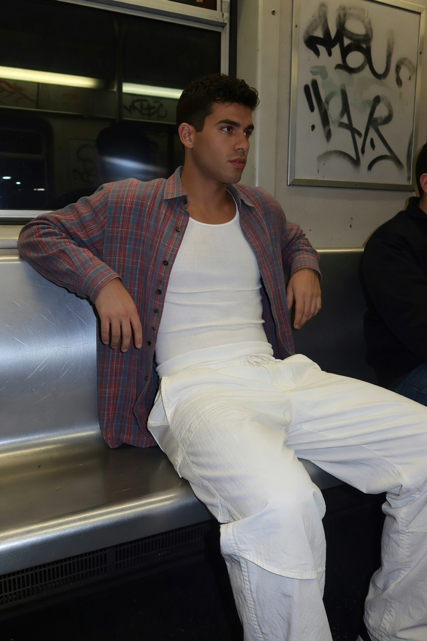 Young man with curly dark hair sits relaxed on a subway bench, wearing an open reddish plaid flannel shirt over a fitted white tank top and loose white parachute pants bunched at the ankles, graffiti-covered walls and another passenger in background