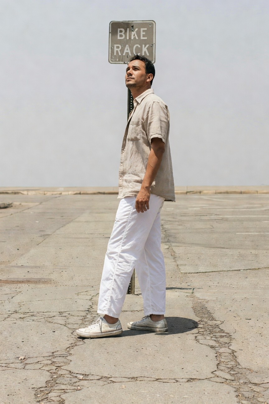 Side view of a man in a beige short-sleeve linen shirt and wide-leg white parachute pants, white sneakers, standing confidently near a bike rack sign in a sunlit parking lot with cracked pavement.