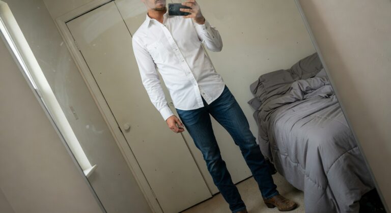 Man taking a mirror selfie in a white open-collar button-up shirt, slim dark blue jeans, and brown leather boots, standing confidently in a bedroom setting