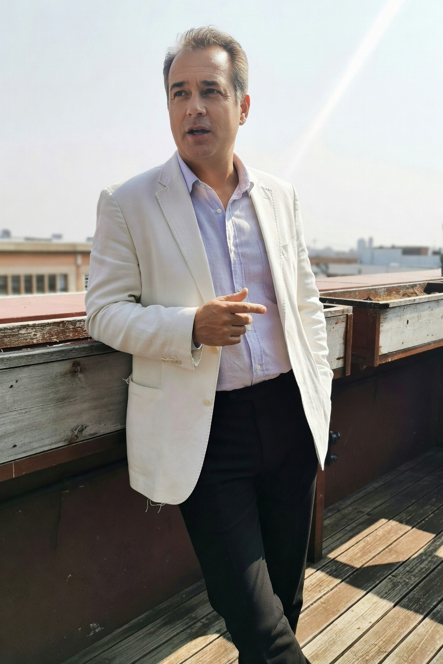 Man in light white blazer over pale blue open-collar shirt and slim black pants poses confidently on urban rooftop pointing outward with city buildings and blue sky in background