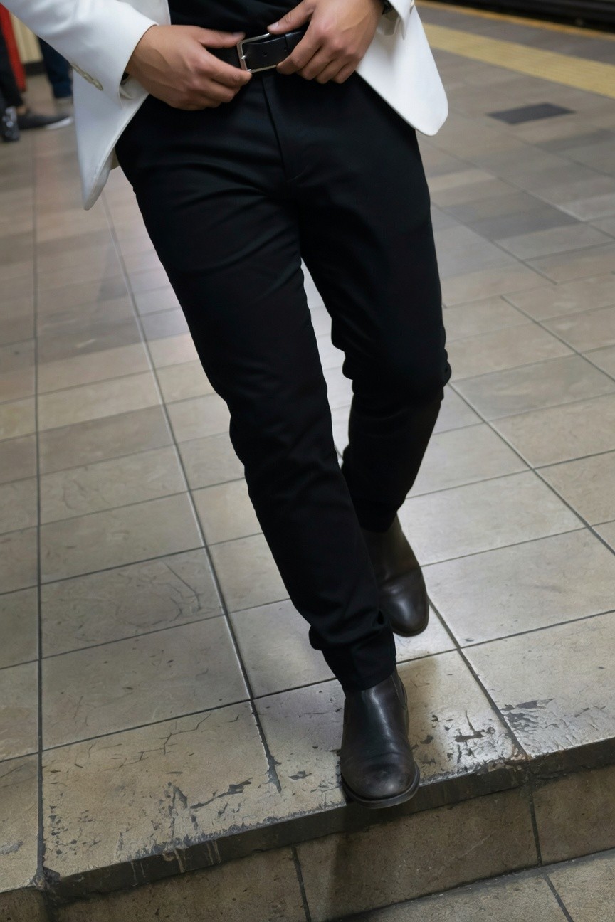 Man in white single-breasted blazer and slim black pants with black Chelsea boots stands confidently on subway platform steps, hand on leather belt buckle, urban tiled background