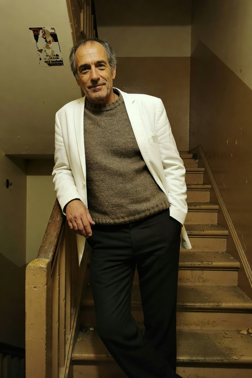 Older man with salt-and-pepper hair smiling confidently on wooden stairs wearing an open white blazer over a chunky brown knit sweater tucked into slim black pants for a refined layered look