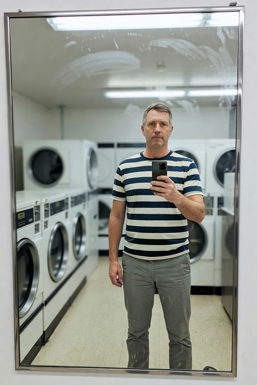 Mirror selfie of a man in a laundromat wearing a short-sleeve navy-and-white striped shirt tucked into slim gray chinos, standing amid rows of white washers and dryers under fluorescent lights
