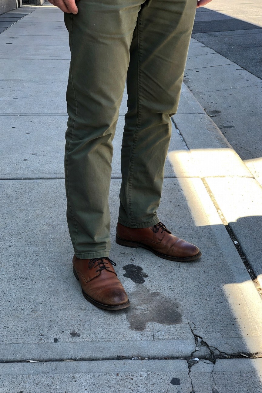 Man in slim-fit olive green chinos and polished brown leather oxford shoes stands confidently on a sunlit urban sidewalk with subtle wear on the pavement.
