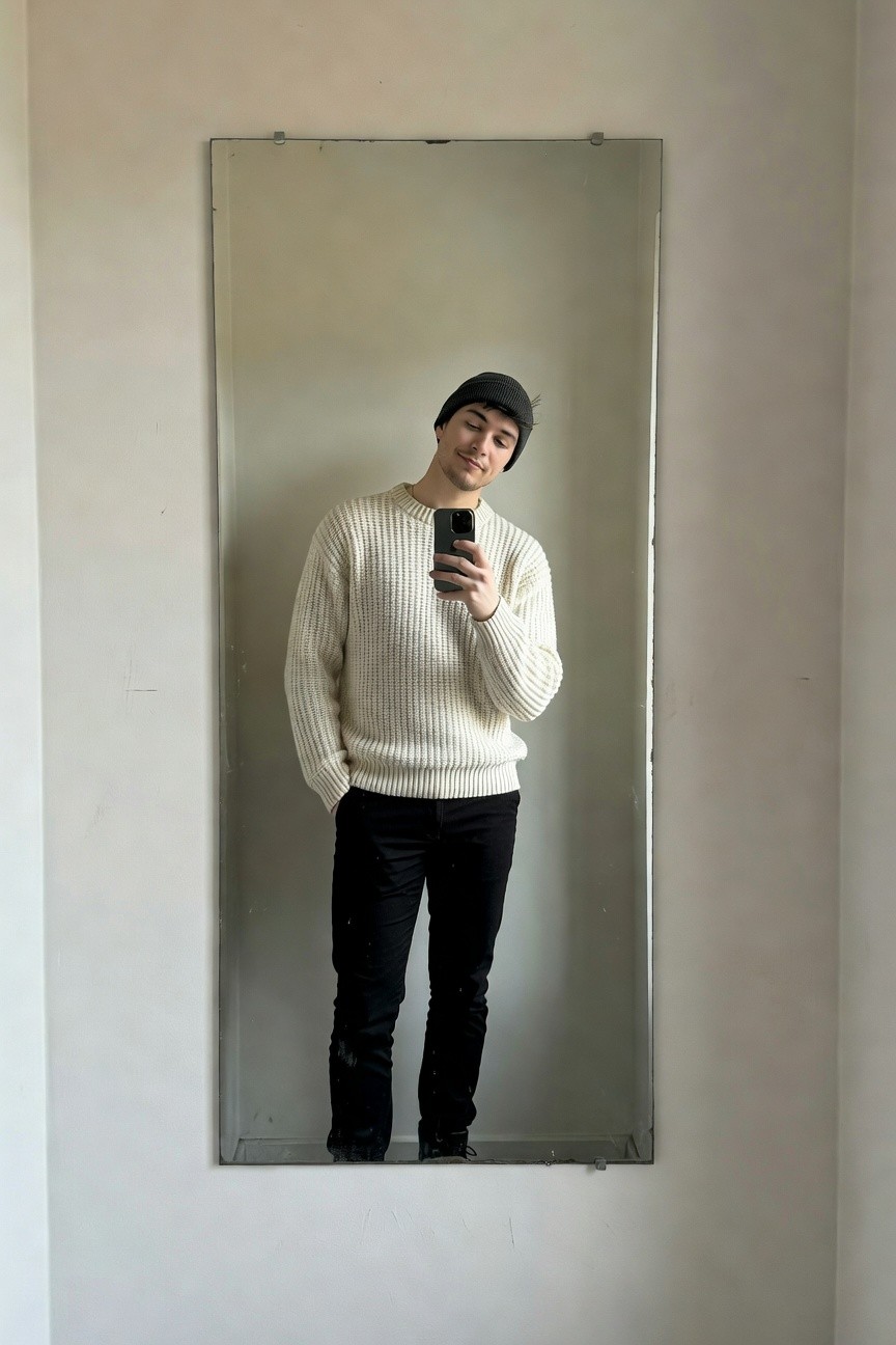 A young man in a white cable-knit sweater, black beanie, and slim black chinos poses in front of a full-length mirror selfie-style, hands in pockets for a casual sharp vibe.