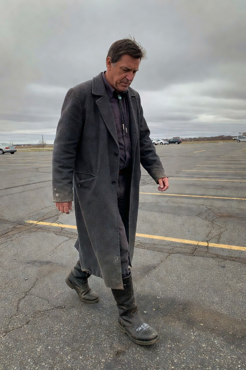 Man in long gray overcoat over dark purple shirt and pants, wearing tall gray boots, walking in parking lot for cowboy aesthetic