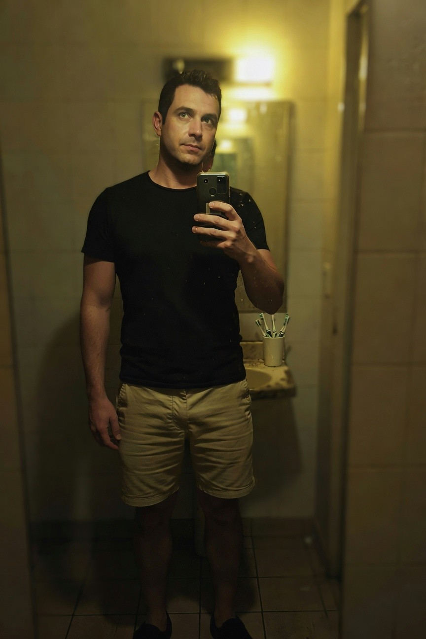 Mirror selfie of a fit man in a yellow-lit bathroom wearing a fitted black short-sleeve t-shirt, mid-length khaki cargo-style shorts, and dark sneakers, standing tall with phone in hand.