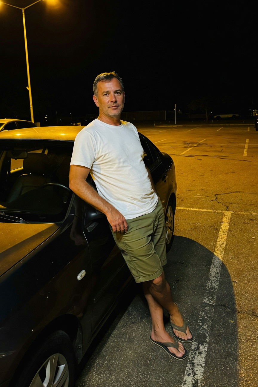 Man in his 40s leaning against a black sedan in a dimly lit parking lot at night, wearing a fitted white cotton T-shirt, olive green knee-length shorts, and black flip-flops, exuding relaxed summer minimalism under streetlamp glow