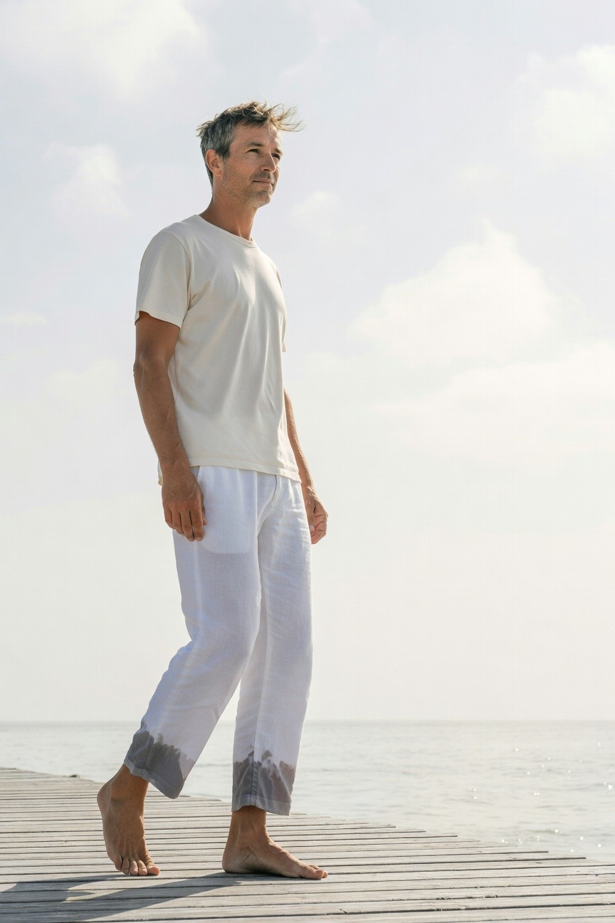 Silver-haired man in cream short-sleeve t-shirt and loose white pants with gray hem fading, walking barefoot on a seaside pier against a hazy ocean backdrop