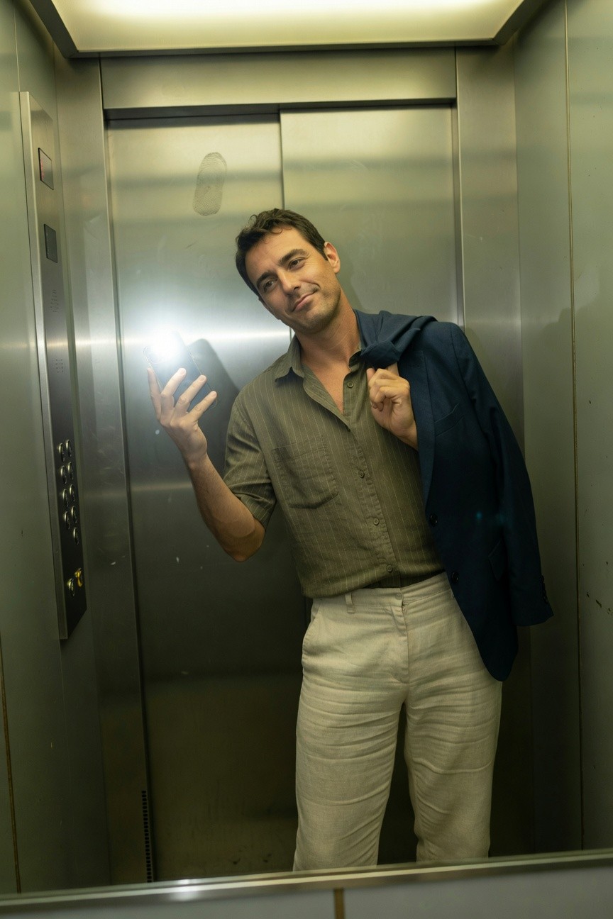 Man taking elevator selfie in olive short-sleeve button-up shirt unbuttoned at collar, navy blazer draped over shoulder, light beige linen trousers, smiling confidently against metallic walls