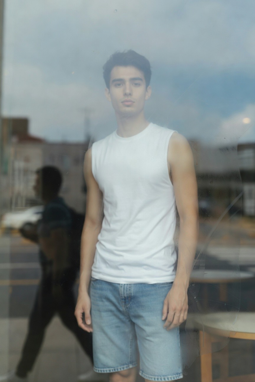 A young man stands casually in front of a foggy window wearing a fitted white sleeveless tank top, light blue denim mid-thigh shorts, and white sneakers, with blurred urban street reflections behind him evoking a serene minimal summer vibe.