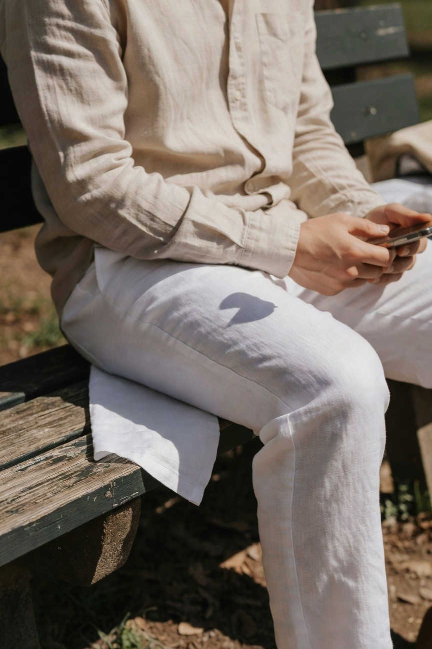 Man in beige linen button-up shirt and white linen pants sits relaxed on outdoor wooden bench, holding phone, sunlight casting bench shadows on pants