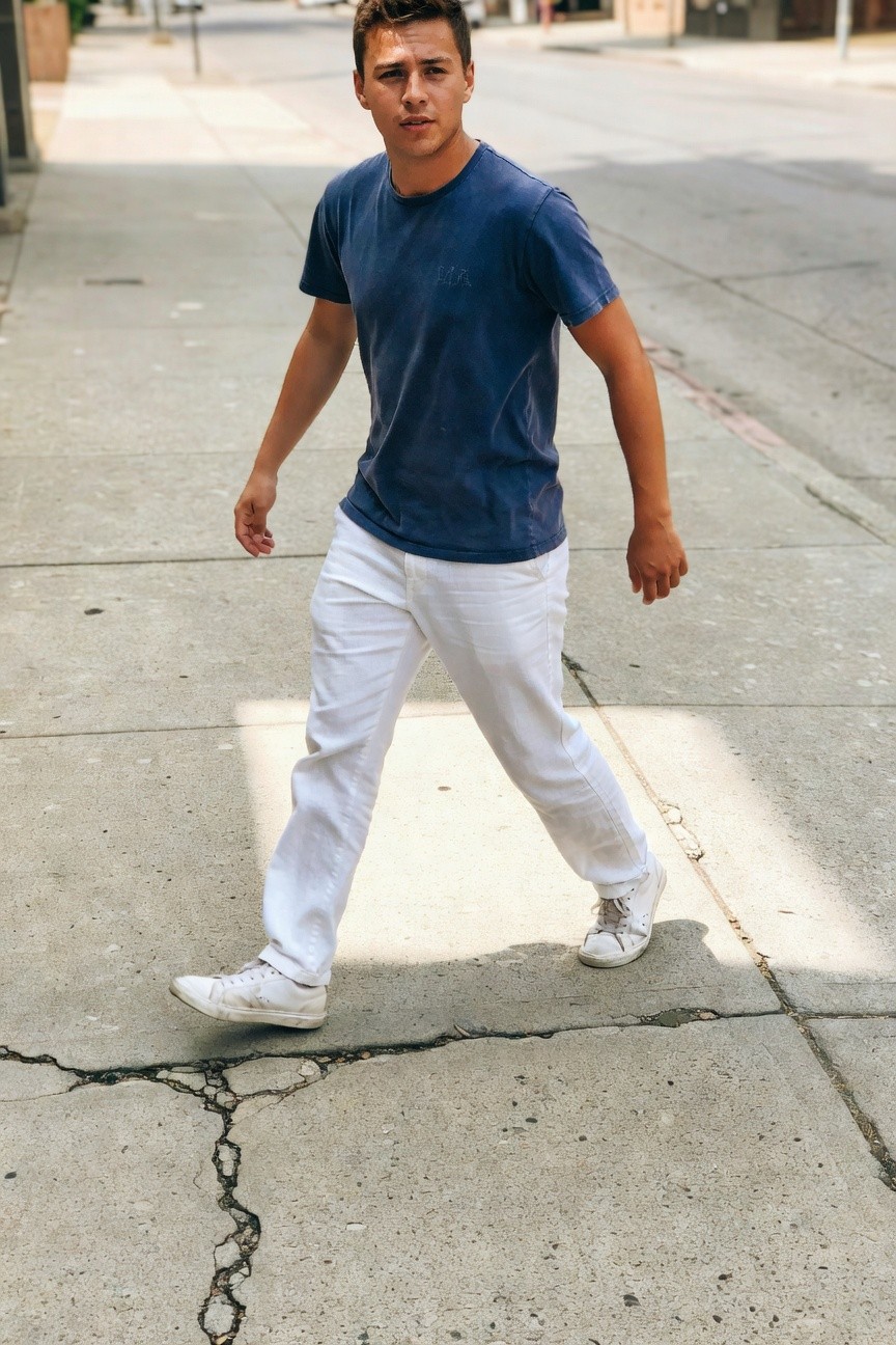 Young man striding on sunny city sidewalk in light blue crewneck t-shirt, loose white linen pants, and white sneakers, shadows stretching long