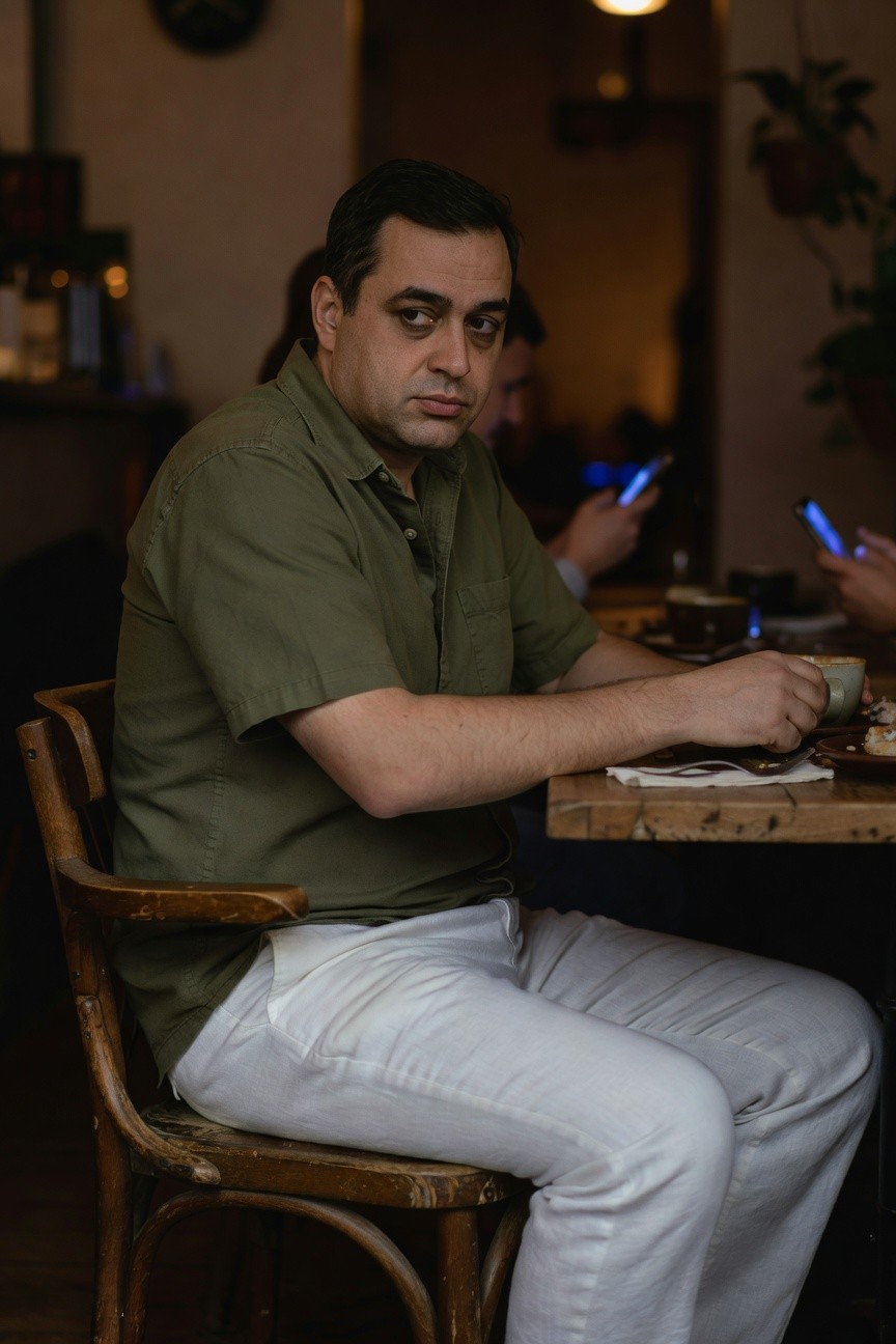 Man in sage green short-sleeved shirt and white wide-leg linen pants sits casually at a rustic cafe table, holding a teacup, with warm ambient lighting and blurred background patrons.
