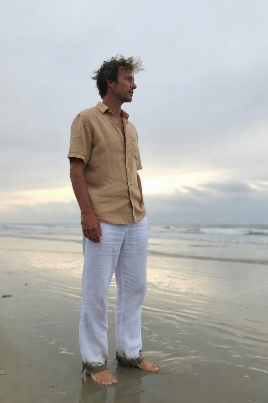A man stands barefoot in shallow ocean water on a beach at dusk, wearing white linen pants rolled at the ankles and a beige short-sleeve linen shirt with open collar, gazing toward the horizon under a cloudy sky.