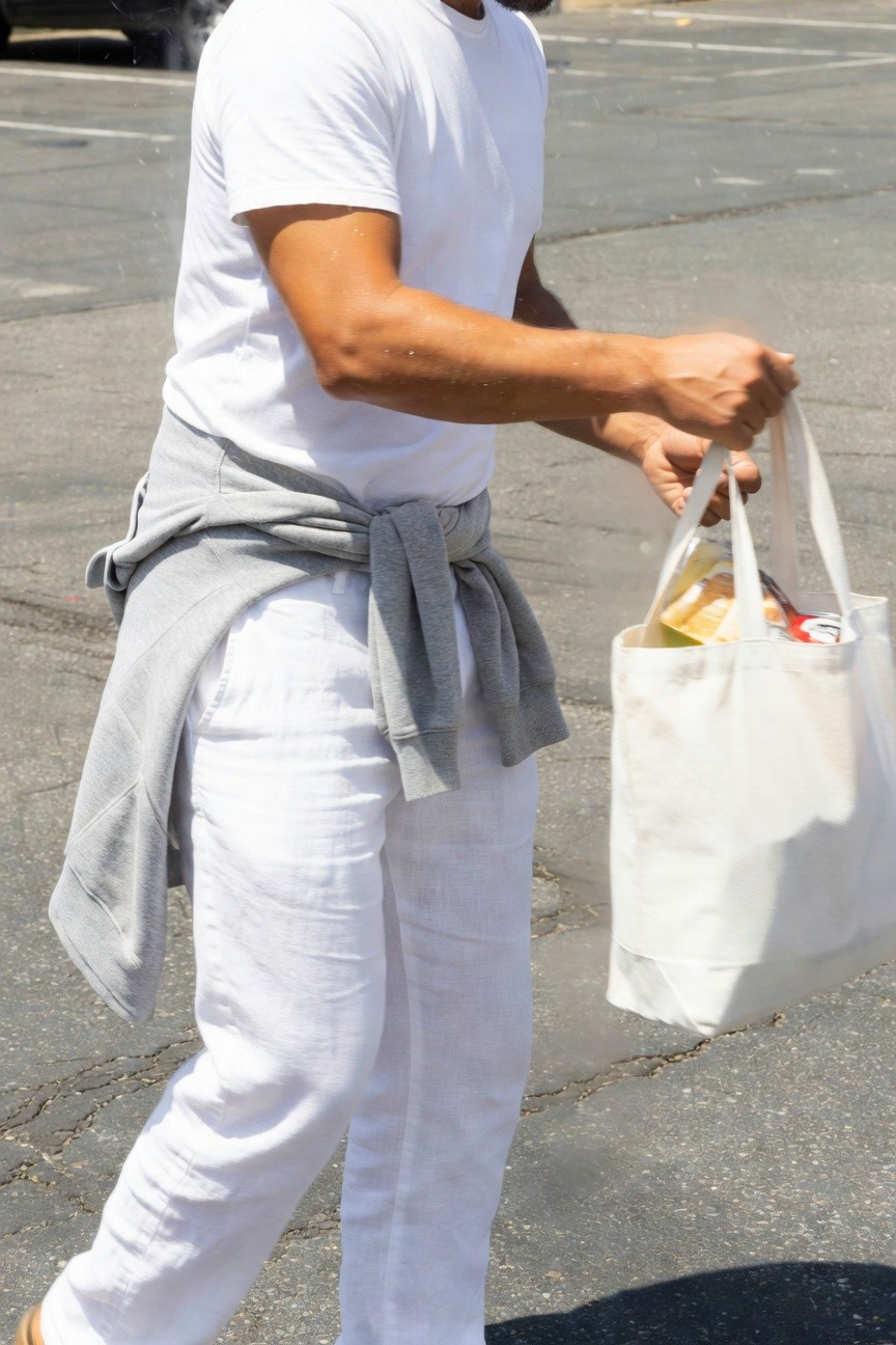 Man in white linen pants and white t-shirt with gray hoodie tied around waist, carrying white canvas tote bag of groceries while walking in sunlit parking lot