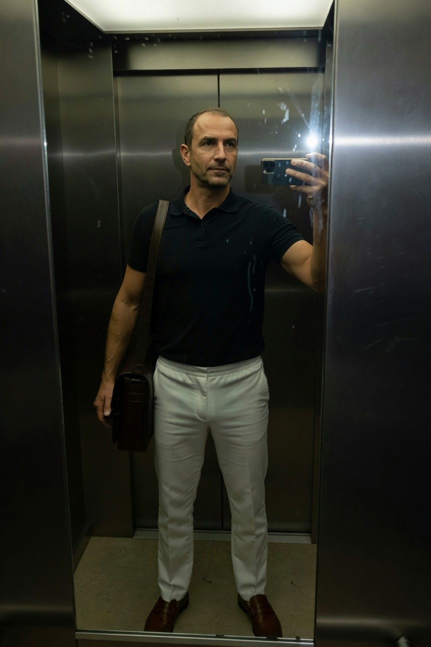 Man in elevator selfie wearing fitted black short-sleeve polo shirt, slim white track pants, brown leather shoulder bag, and matching brown leather shoes, with reflective metallic walls and overhead light.