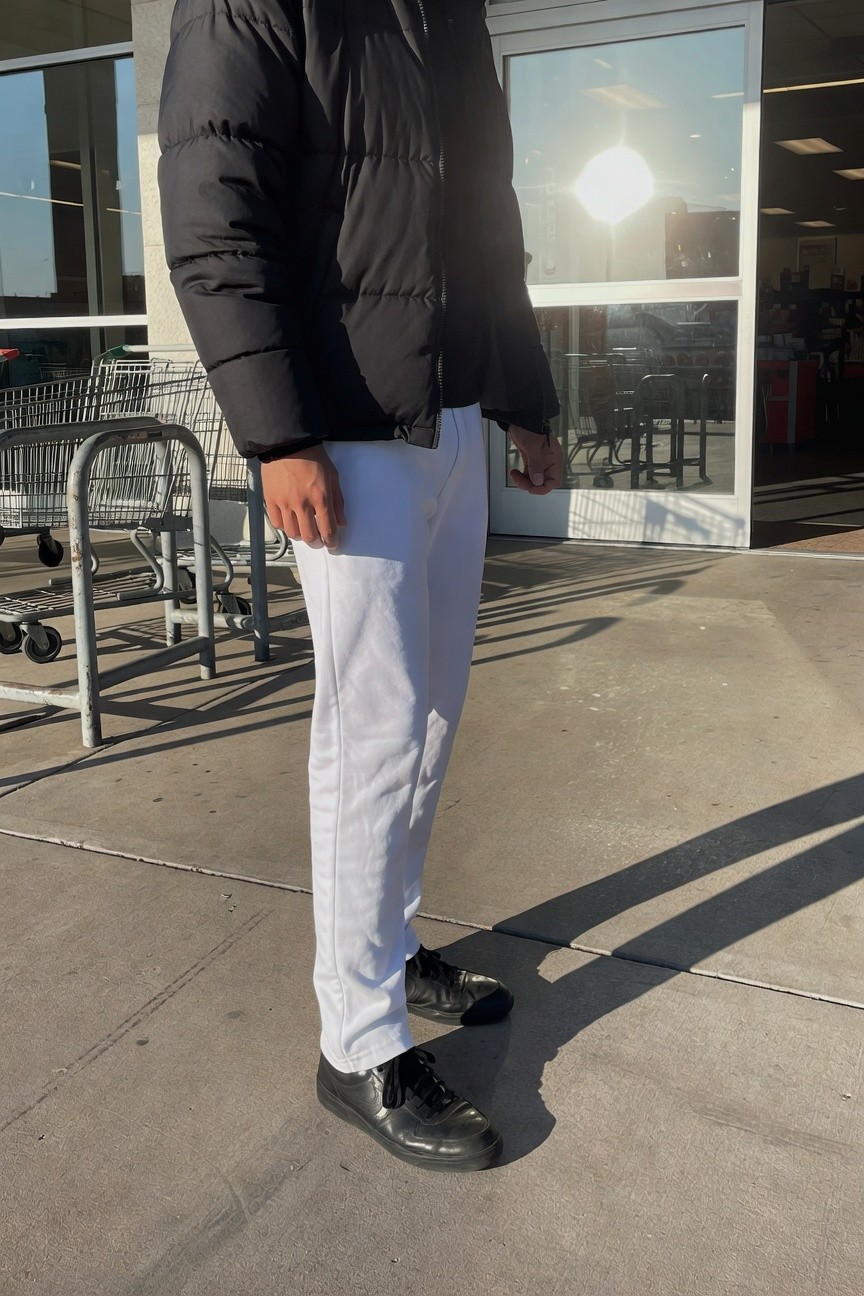 Man in oversized shiny black puffer jacket, slim white track pants with black side stripes, and chunky black sneakers stands casually outside a big-box store entrance, sunlight bouncing off glass doors and pavement