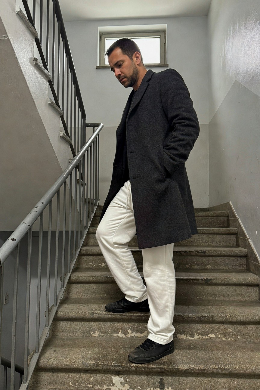 A man with short dark hair stands mid-stride on concrete stairs in a dimly lit stairwell, wearing a long charcoal wool overcoat over a black turtleneck, loose white track pants, and black sneakers, one hand in pocket, gazing downward thoughtfully.