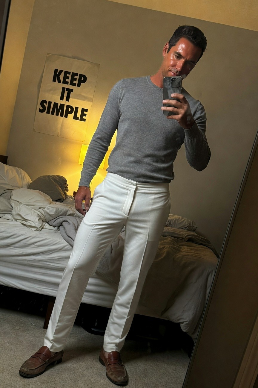 Man in slim-fit grey crewneck sweater, white tailored track pants, and brown loafers, mirror selfie in cozy bedroom with KEEP IT SIMPLE wall art and soft lamp light