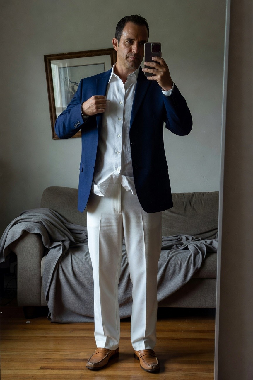 Man in navy blazer over unbuttoned white dress shirt and slim white track pants, brown loafers, selfie in cozy living room with couch and mirror