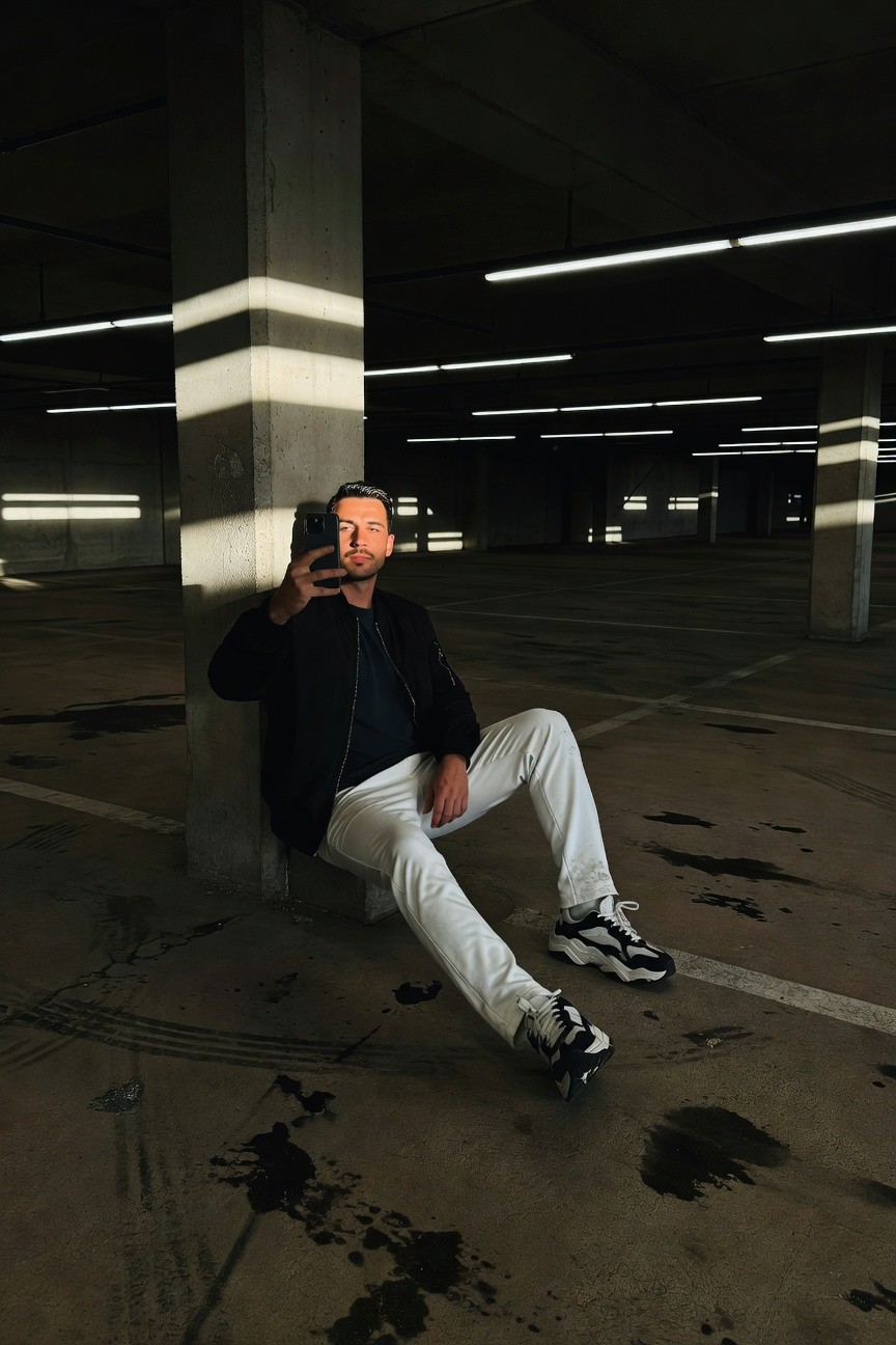 Man in black bomber jacket and white track pants sits casually against a concrete pillar in an empty parking garage, chunky white sneakers with black accents, dramatic fluorescent lighting casting long shadows