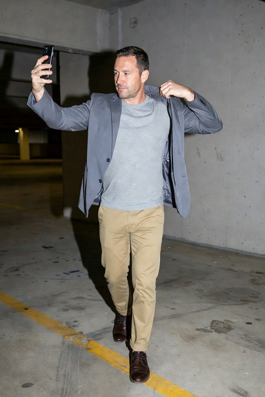 Man in open light gray blazer over gray t-shirt, khaki pants, and brown dress shoes, posing confidently with selfie in underground parking garage
