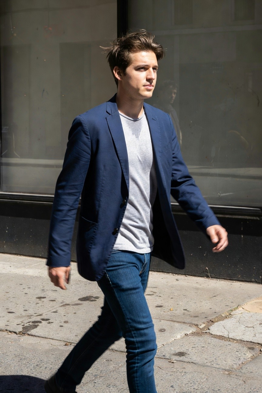Young man in slim-fit navy blazer over white t-shirt and slim blue jeans walking confidently on sunny urban sidewalk, modern business casual style