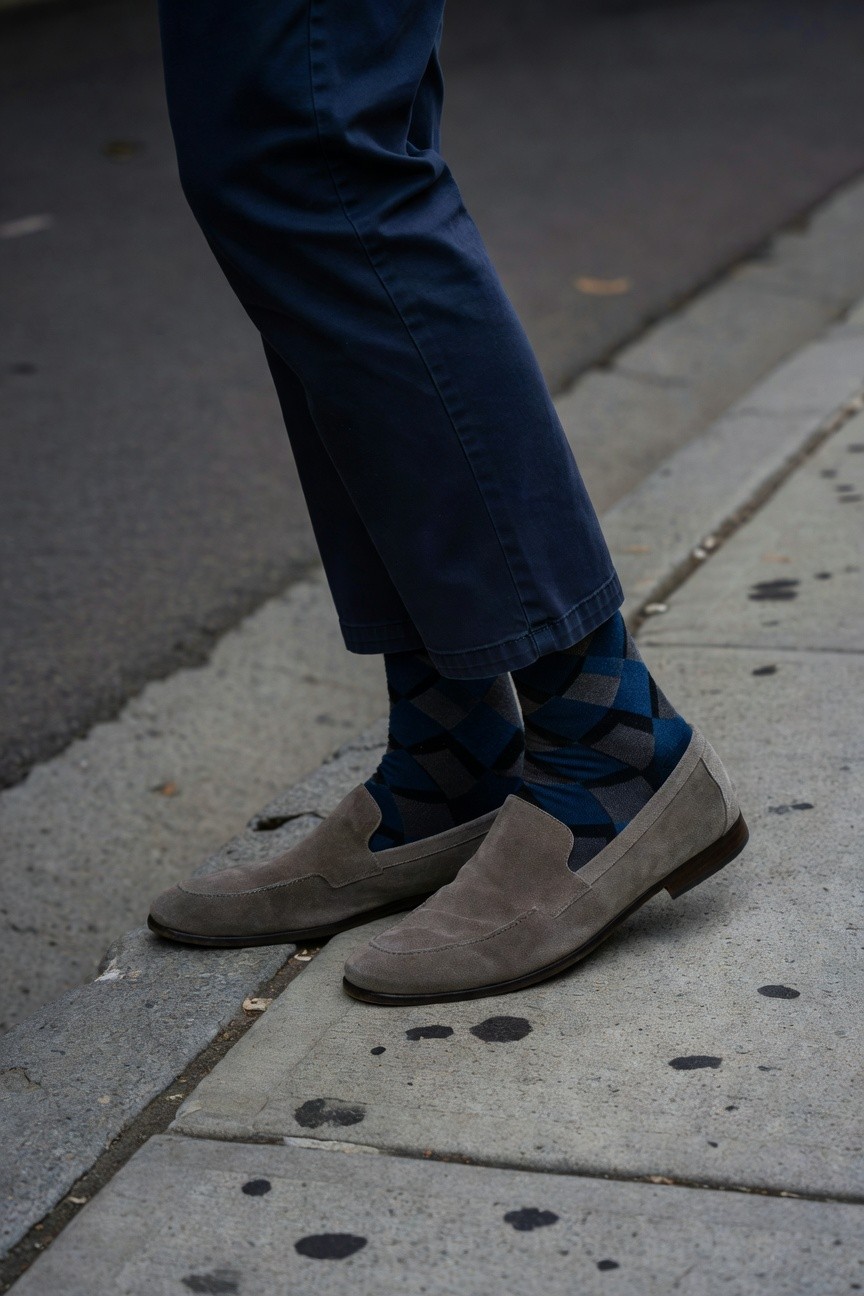 Close-up street shot of a man's lower legs in slim navy chinos cuffed at ankles revealing blue-gray argyle socks and pale gray suede loafers on urban pavement