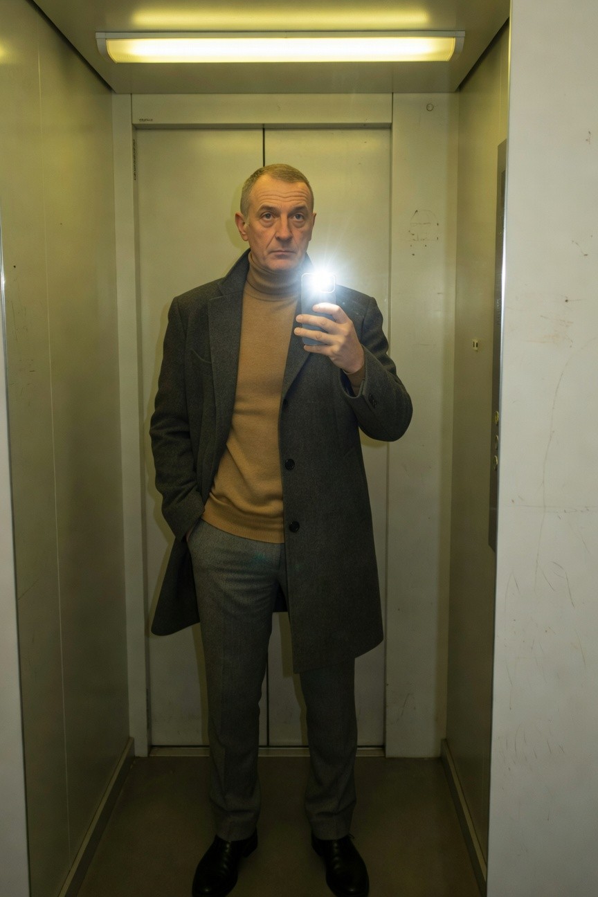 Man in elevator taking selfie wearing open dark grey wool overcoat tan turtleneck light grey slim trousers and black dress shoes neutral tones polished business casual vibe