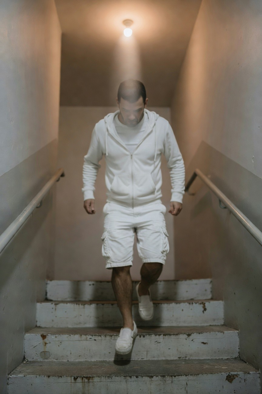 Man in all-white zip-up hoodie, matching cargo shorts, and sneakers descending worn wooden stairs in a dimly lit narrow hallway with beige walls and railing, dramatic overhead light casting soft shadows