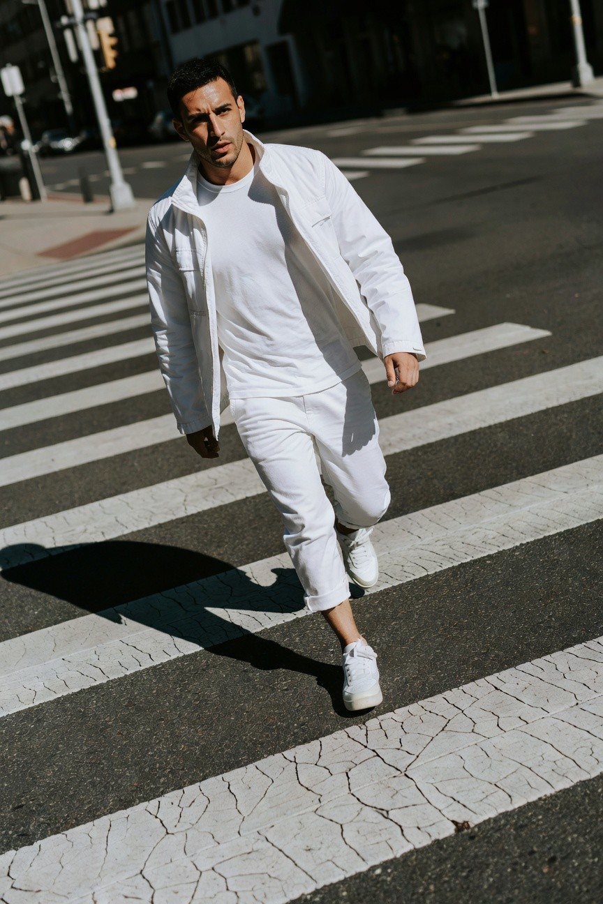 A handsome man with short dark hair crosses a sunlit urban zebra crossing in a chic all-white outfit, featuring an open lightweight white chore jacket over a fitted white t-shirt, cropped white pants, and crisp white sneakers, with city buildings and traffic in the background.