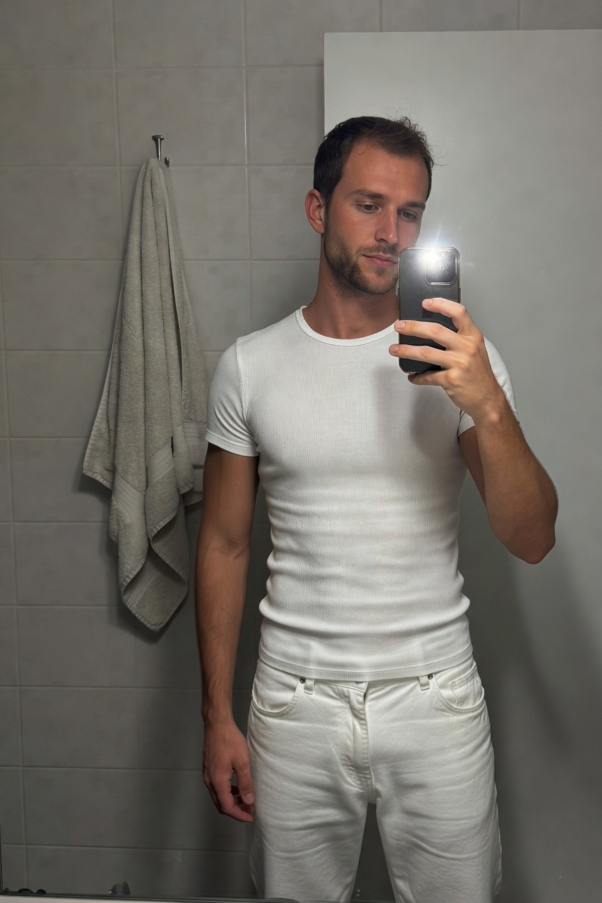 Man taking a mirror selfie in a tiled bathroom wearing a fitted white short-sleeve t-shirt and slim white pants, towel hanging nearby, phone flashlight illuminating the all-white outfit