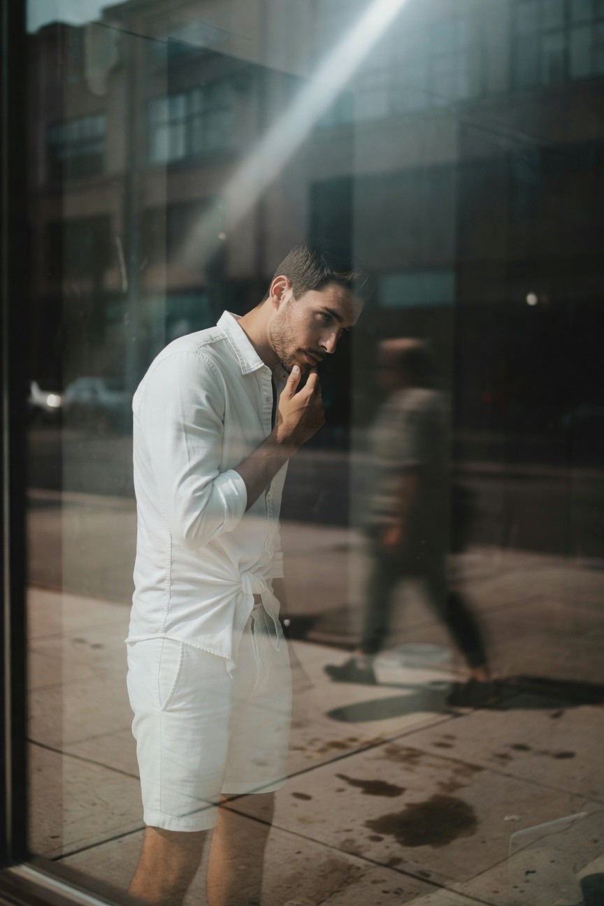 A man in a white linen open shirt and matching shorts stands thoughtfully by a sunlit window reflection on a city street, cigarette in hand, evoking relaxed summer chic.