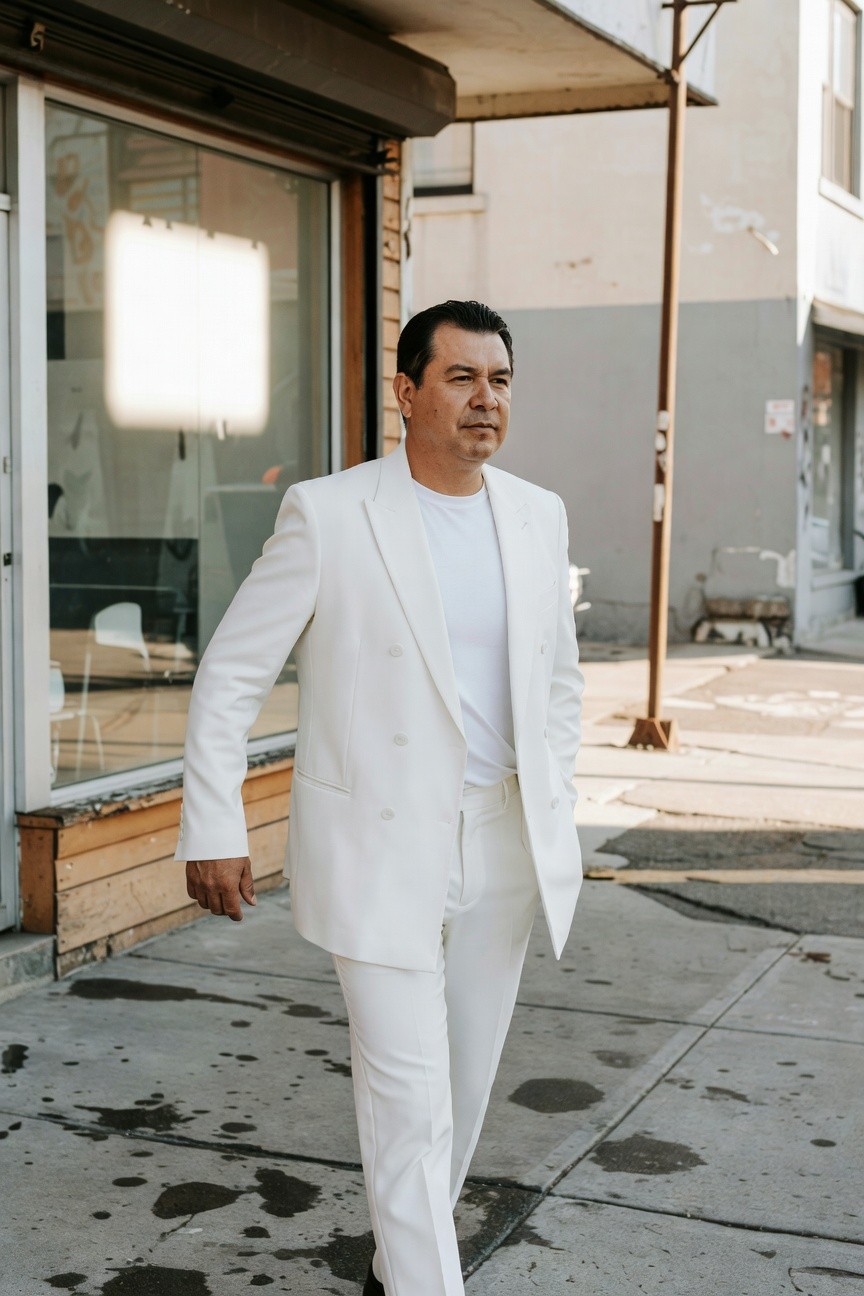 Man in tailored all-white suit jacket, white shirt, and trousers walking confidently on a sunlit urban sidewalk near a storefront, evoking sharp summer sophistication.