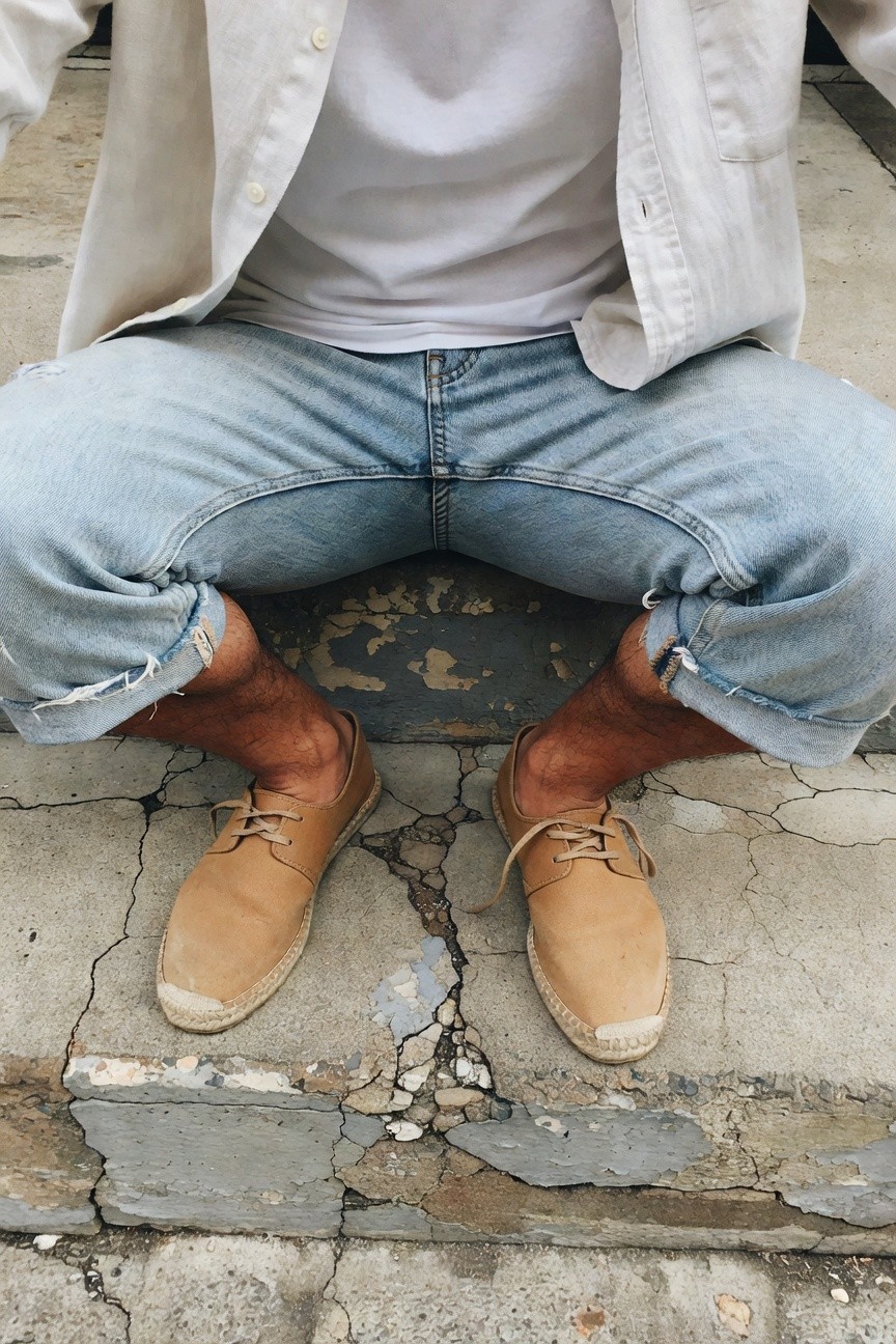 A man sits casually on weathered concrete steps, dressed in an open cream linen jacket over a plain white t-shirt, light blue jeans with rolled cuffs exposing ankles, and tan espadrilles with rope soles, evoking a laid-back summer style against a textured gray background