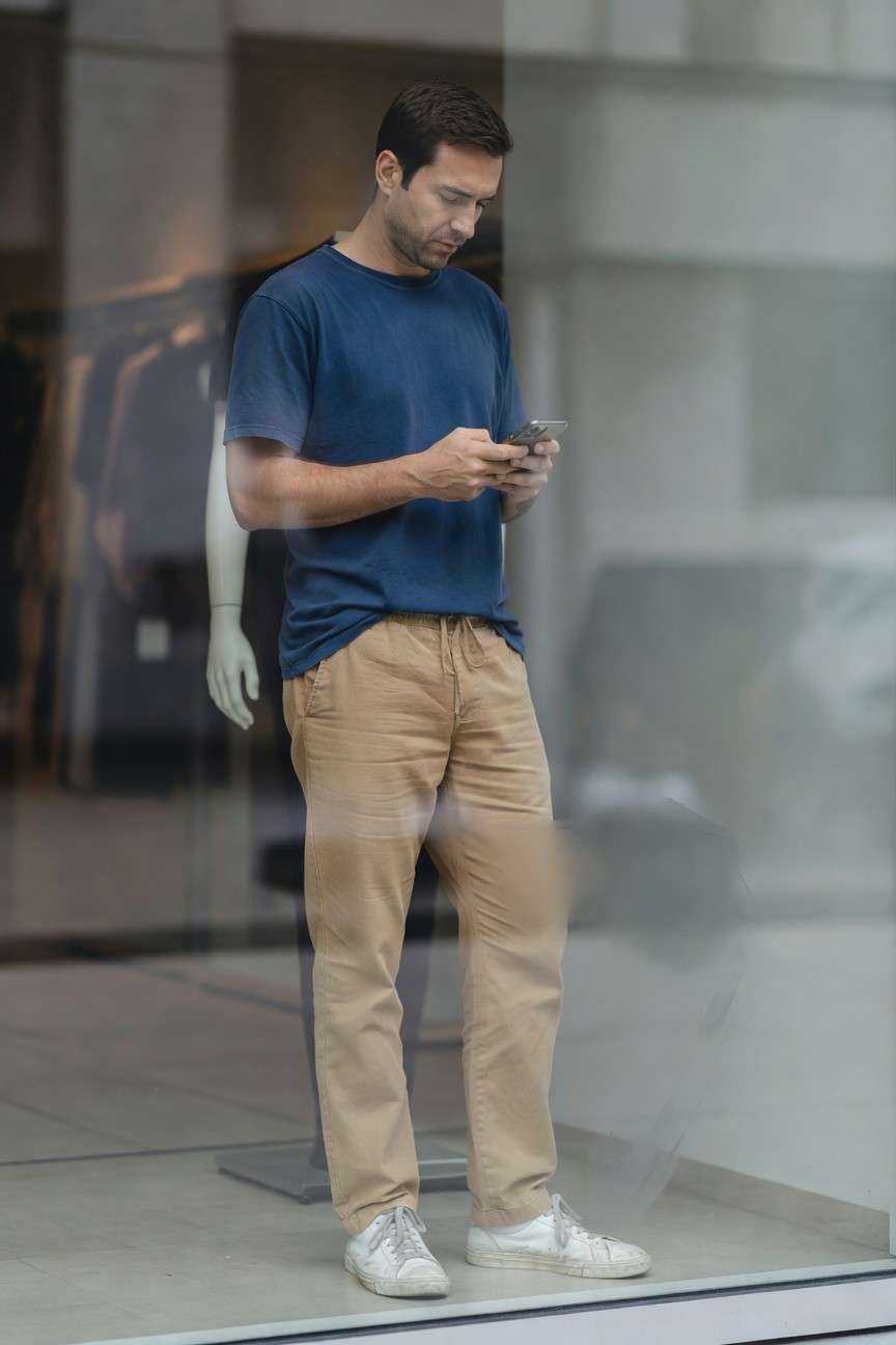 A man stands casually outside a modern store window wearing a fitted navy blue t-shirt, loose khaki drawstring pants, and white sneakers, holding his phone while surrounded by clothing displays inside