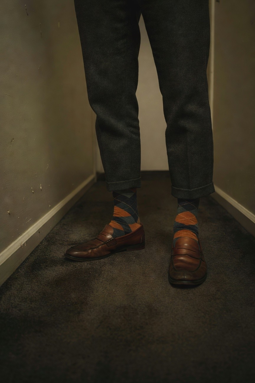 Close-up of a man’s lower legs and feet in a dimly lit hallway, wearing slim gray wool trousers cuffed at the ankles, colorful argyle socks in navy, orange, and gray, and shiny brown leather loafers on a dark carpet floor