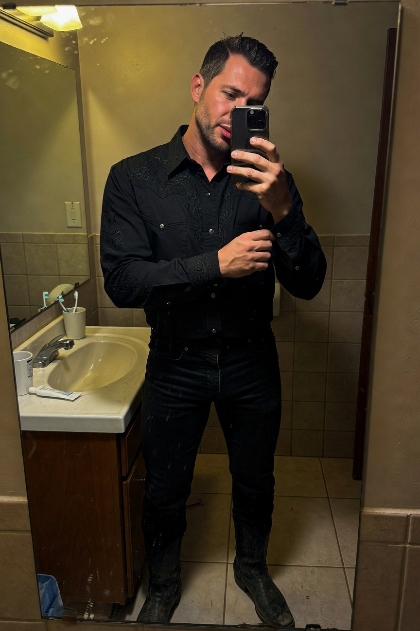 Mirror selfie of a man in a fitted black long-sleeve button-down shirt tucked into slim black jeans and black cowboy boots, standing in a tiled bathroom with sink visible.