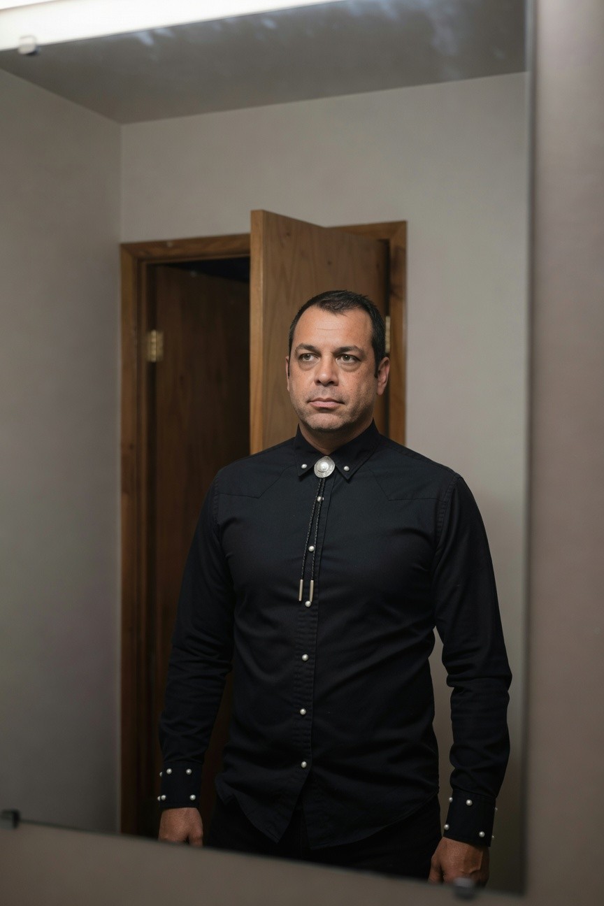 Man standing in front of mirror wearing black snap button shirt with silver-tipped bolo tie, long sleeves cuffed, in indoor setting