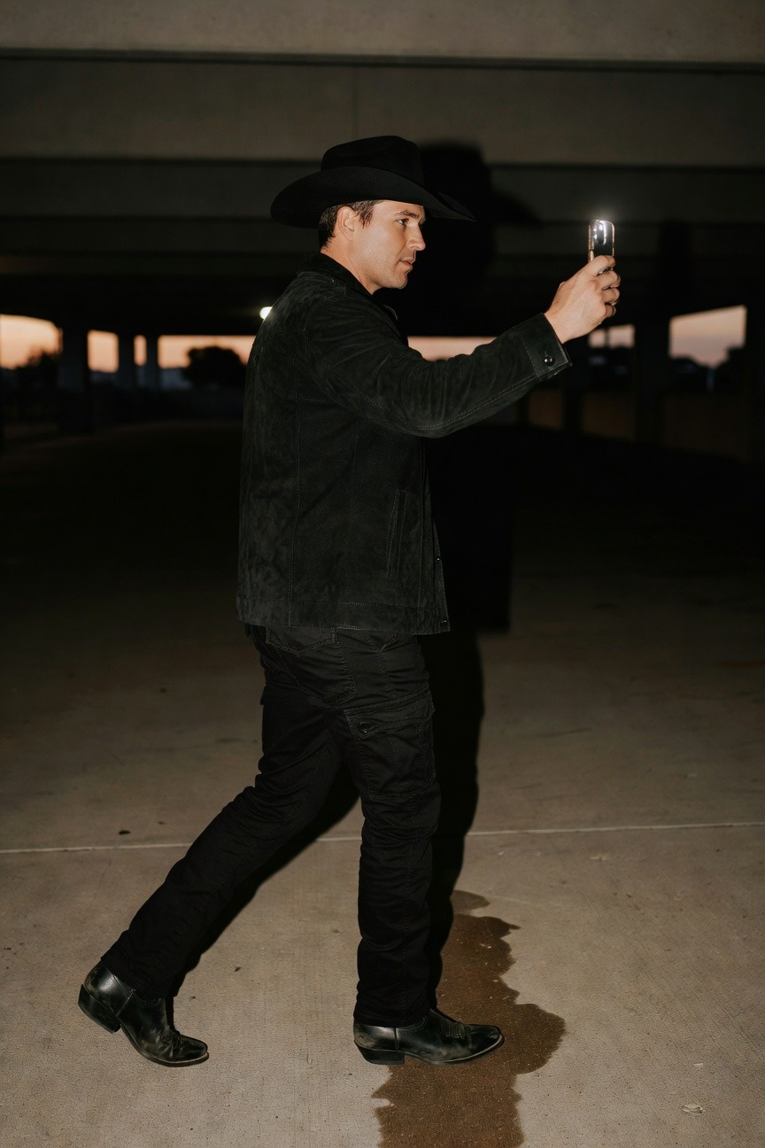 Man in all-black cowboy outfit taking a selfie: black cowboy hat, suede jacket, slim pants, and cowboy boots under a parking structure